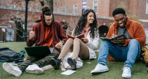 Three university students sit on a grass bank together. They are smiling as they study.